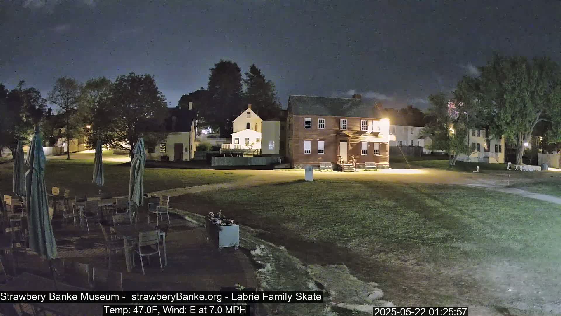 A nighttime view of a grassy area with several historic-looking buildings and outdoor patio furniture under a partly cloudy sky.