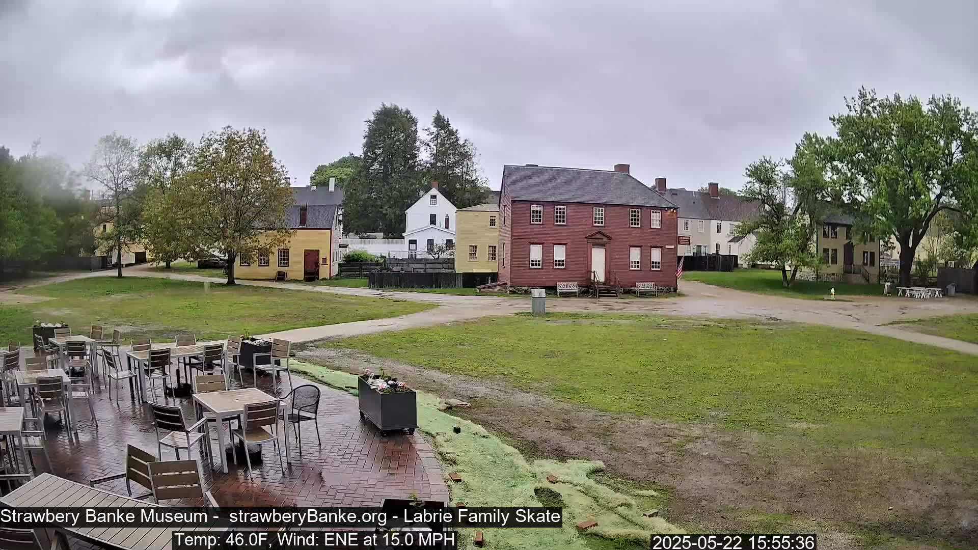 A brick patio with empty tables and chairs overlooks a grassy area and several historic-looking buildings on an overcast day.