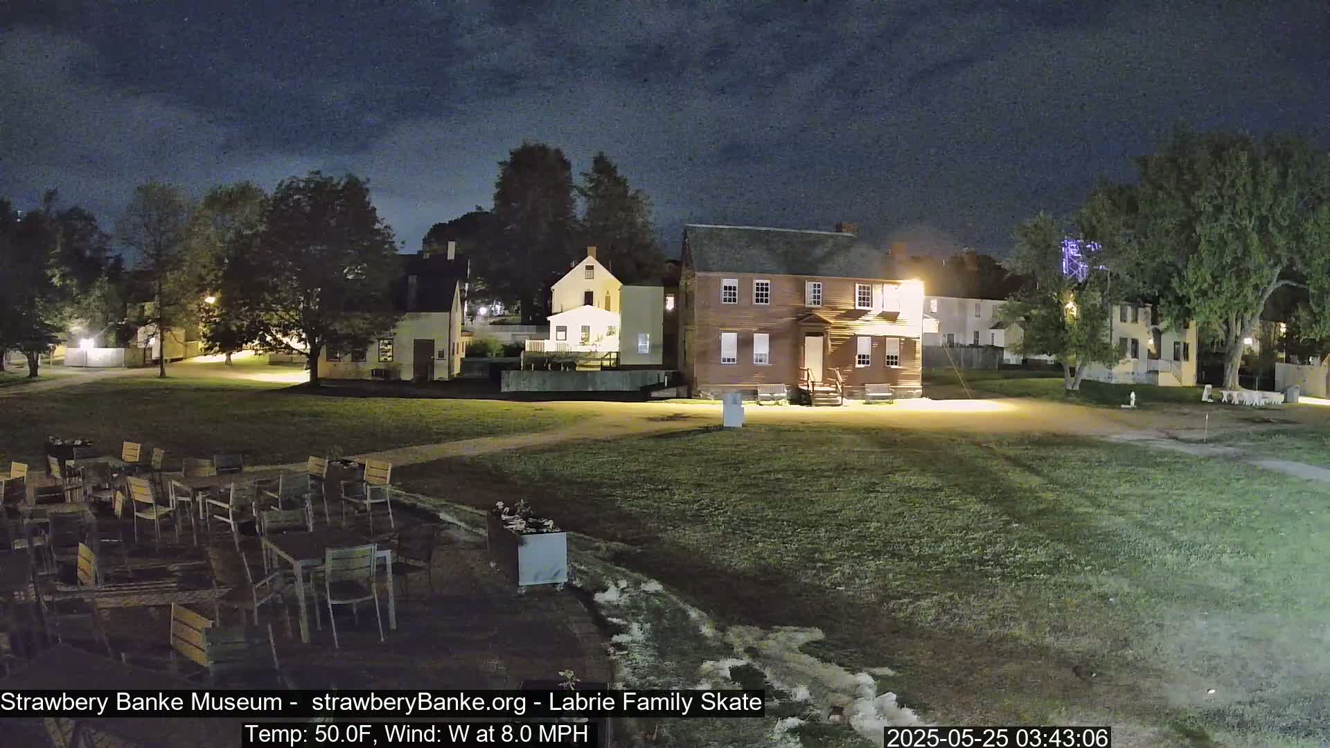 At night, under a dark sky, several colonial-style buildings sit on a grassy area with outdoor seating, and an eight-mile-per-hour westerly wind at 50°F.