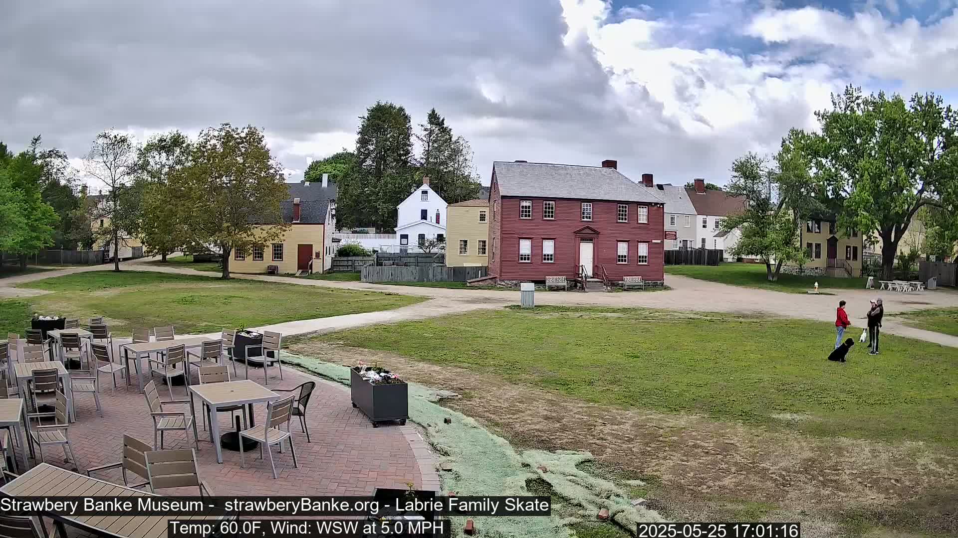 A patio with outdoor furniture overlooks a grassy area with several historic-looking buildings under a partly cloudy sky.