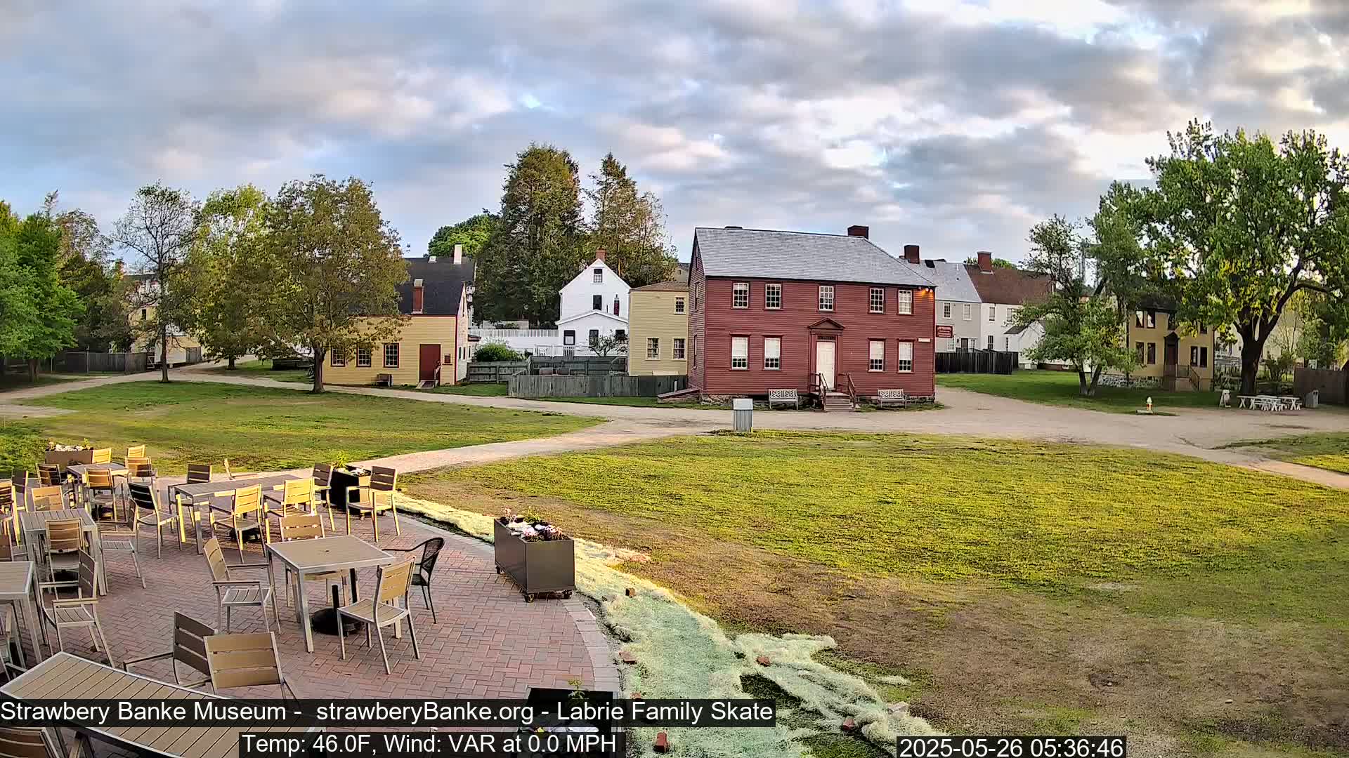 A patio with outdoor furniture sits in front of a grassy area and several historic buildings under a partly cloudy sky.