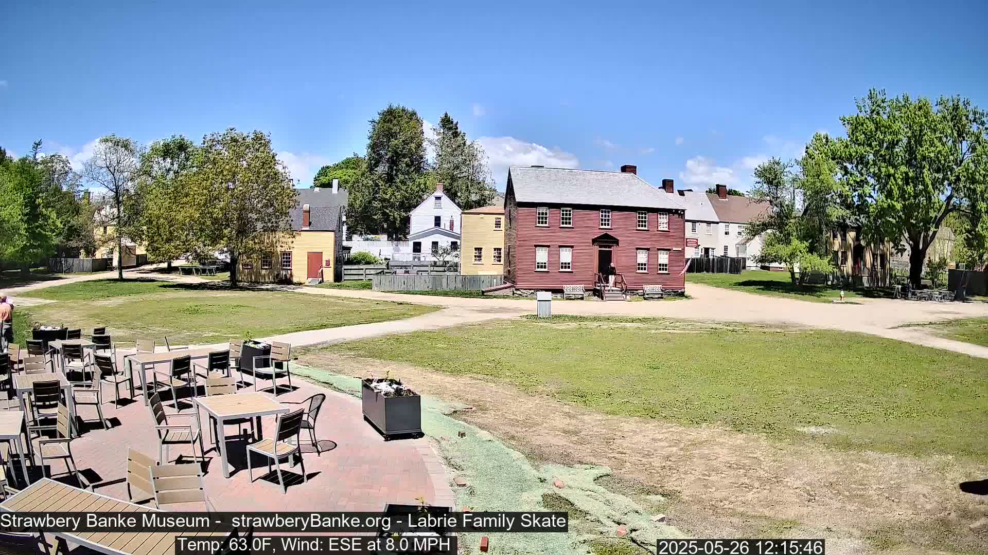 A sunny outdoor scene shows a courtyard with several tables and chairs, situated near several historic-looking buildings under a clear blue sky.