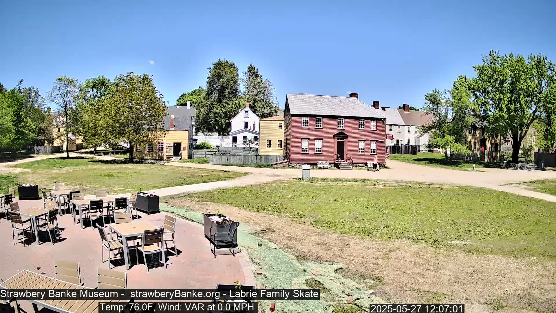 A sunny outdoor scene shows several colonial-style buildings surrounding a grassy area with an outdoor patio containing several tables and chairs.