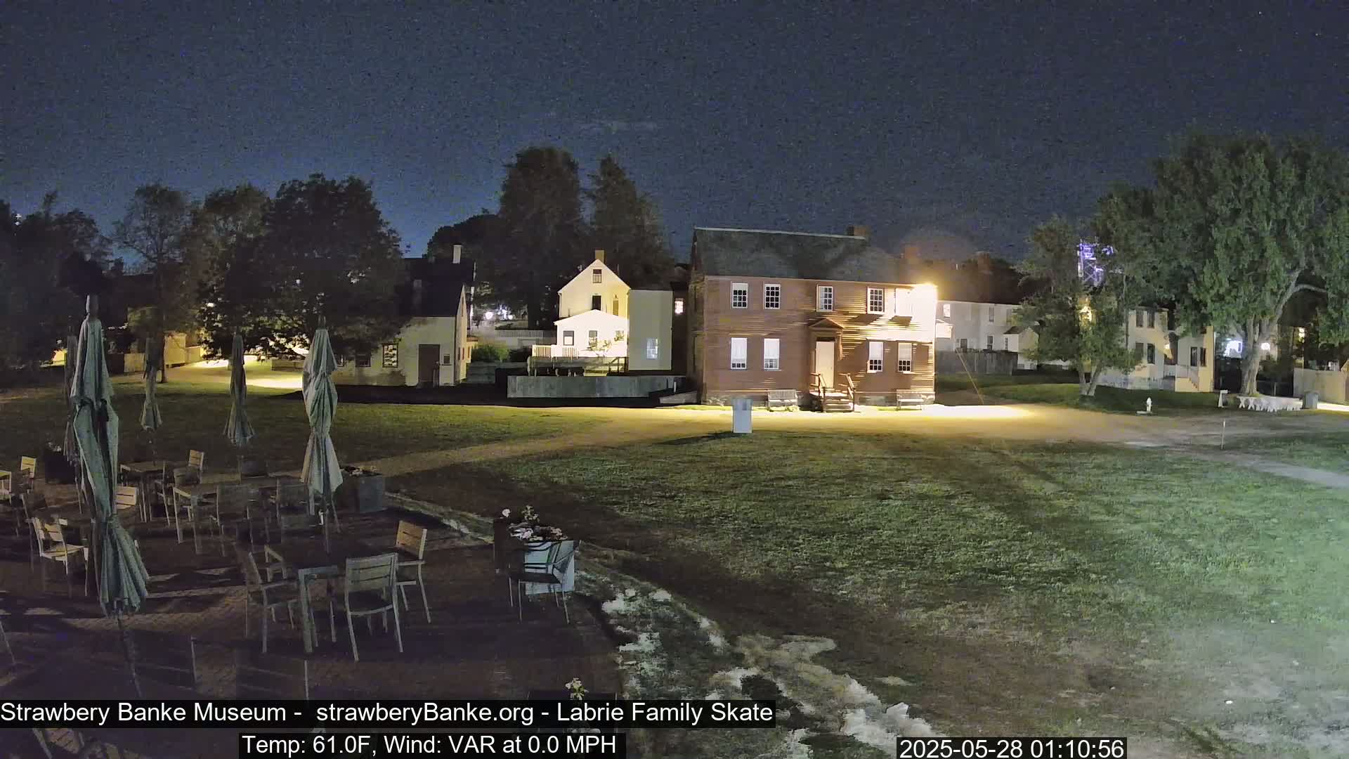 An outdoor nighttime scene shows several historical-looking buildings illuminated, a grassy area with patio furniture, and calm weather conditions.