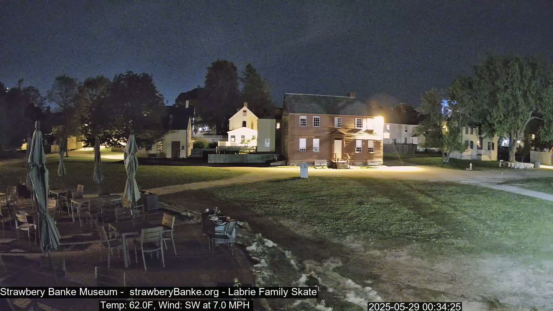 A nighttime outdoor scene shows a grassy area with several tables and chairs under umbrellas, situated in front of several historic-looking buildings under a dark, clear sky.