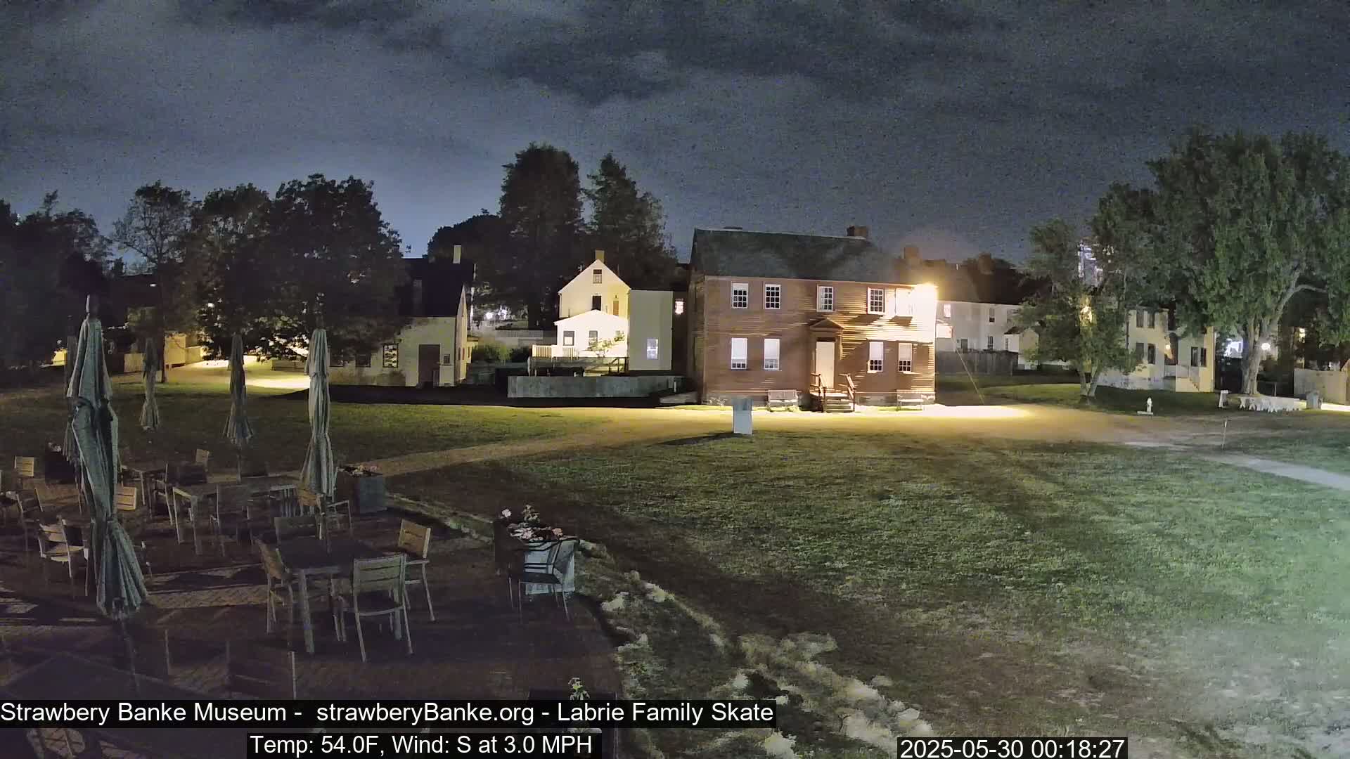 A nighttime view of a grassy area with several historic-looking buildings, outdoor seating with umbrellas, and trees under a partly cloudy sky.