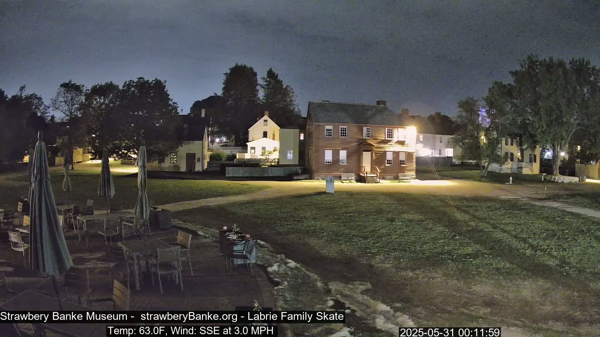 A nighttime view of a grassy area with several illuminated historical buildings, outdoor patio furniture, and umbrellas under a dark sky with a gentle SSE wind.