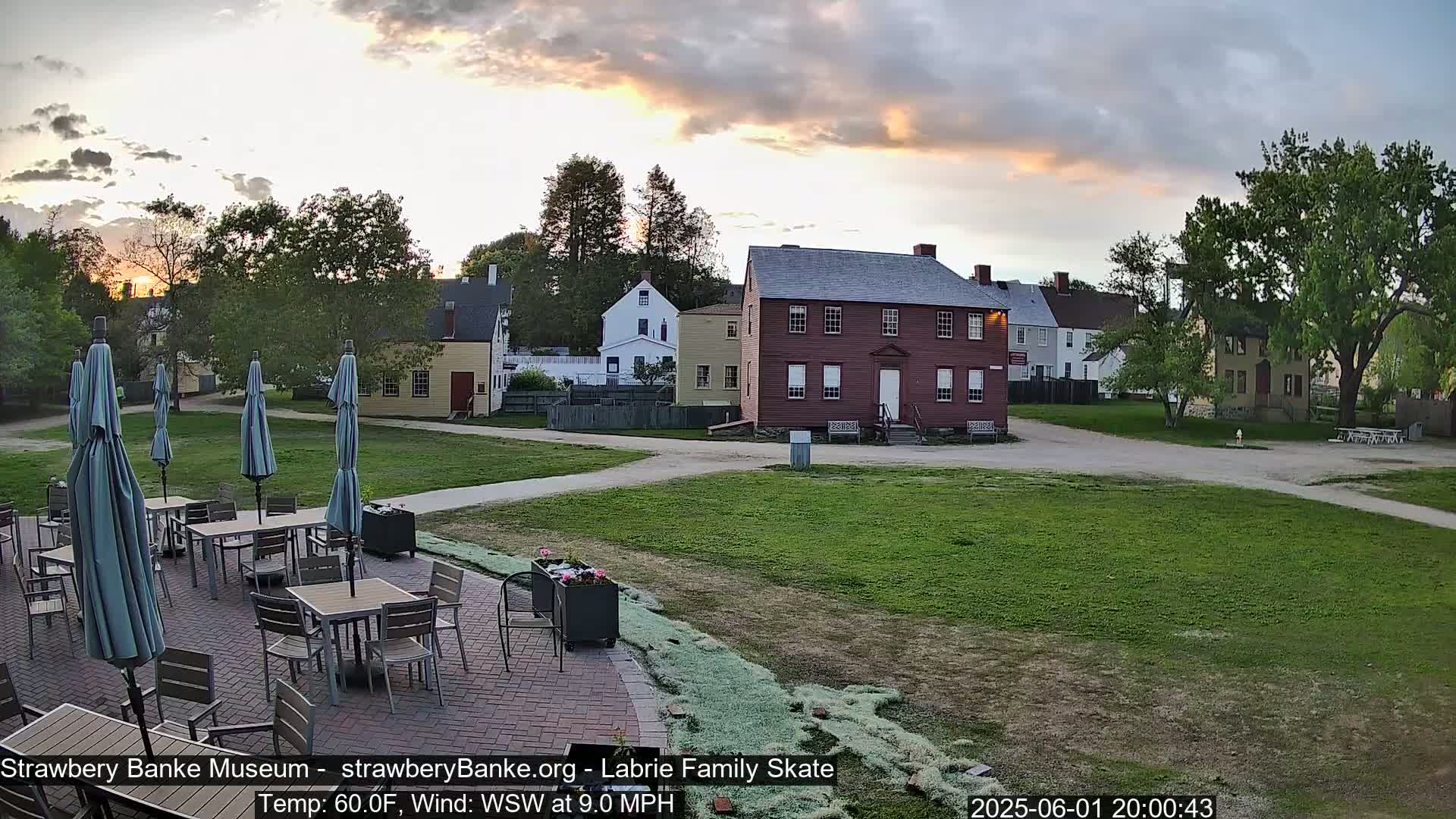 A patio with tables and umbrellas sits adjacent to a grassy area overlooking several historic-looking buildings at sunset under a partly cloudy sky.