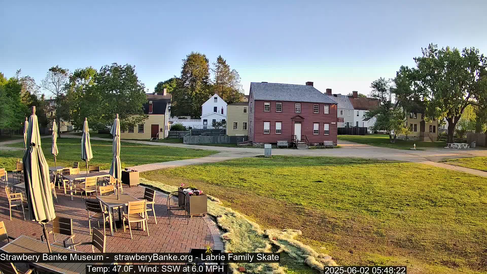 A brick patio with tables and umbrellas sits before several historic-looking buildings on a grassy lawn, under a clear sky with a light wind.