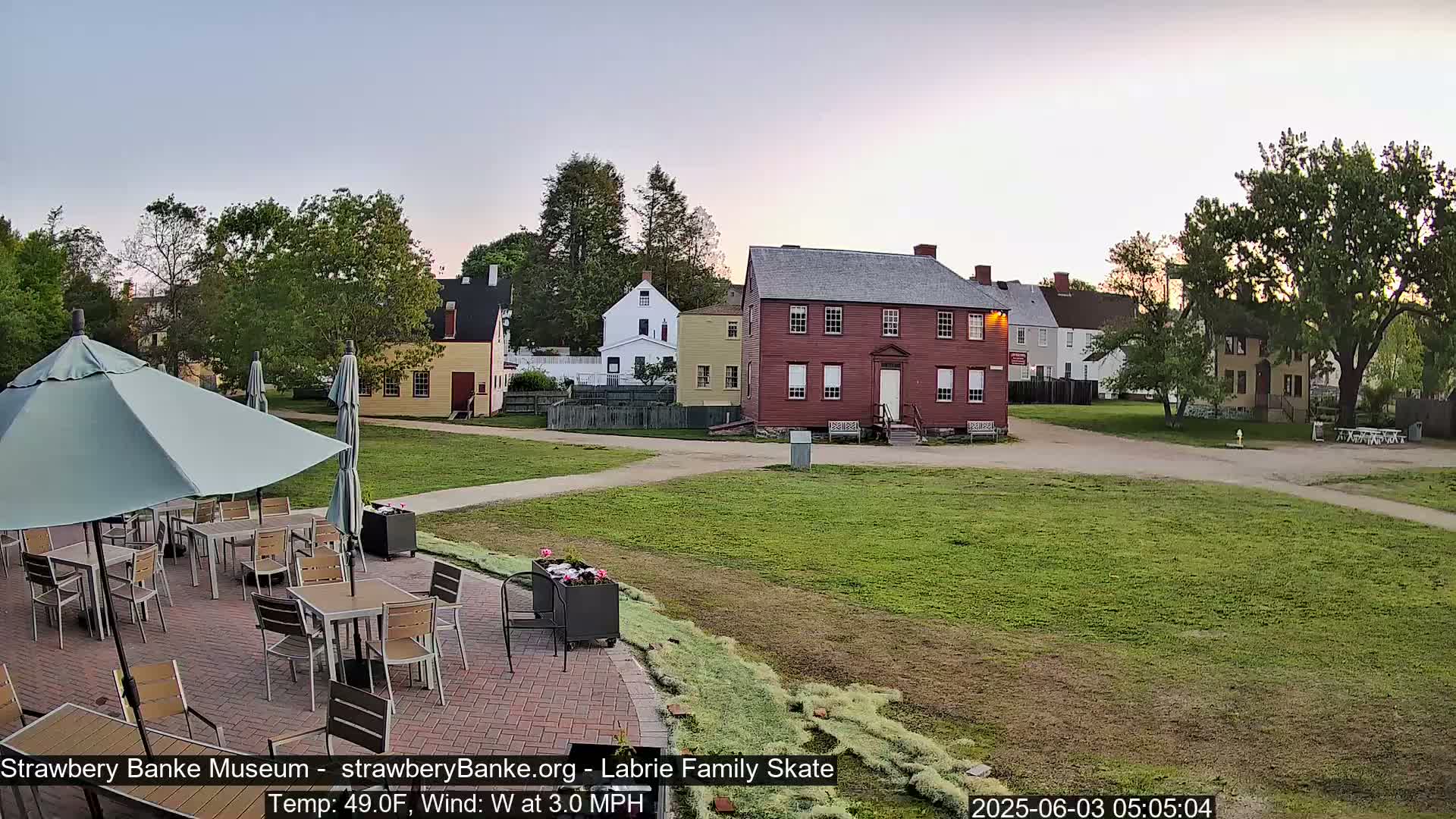 An outdoor patio with tables and umbrellas sits before a grassy area and several historic-looking buildings under a clear, twilight sky.