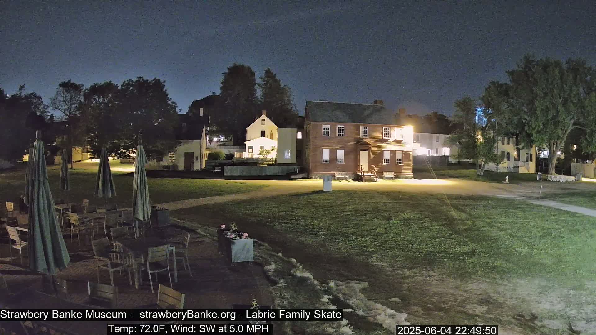 A nighttime scene shows several colonial-style buildings and a grassy area with outdoor tables and umbrellas under a clear, dark sky.