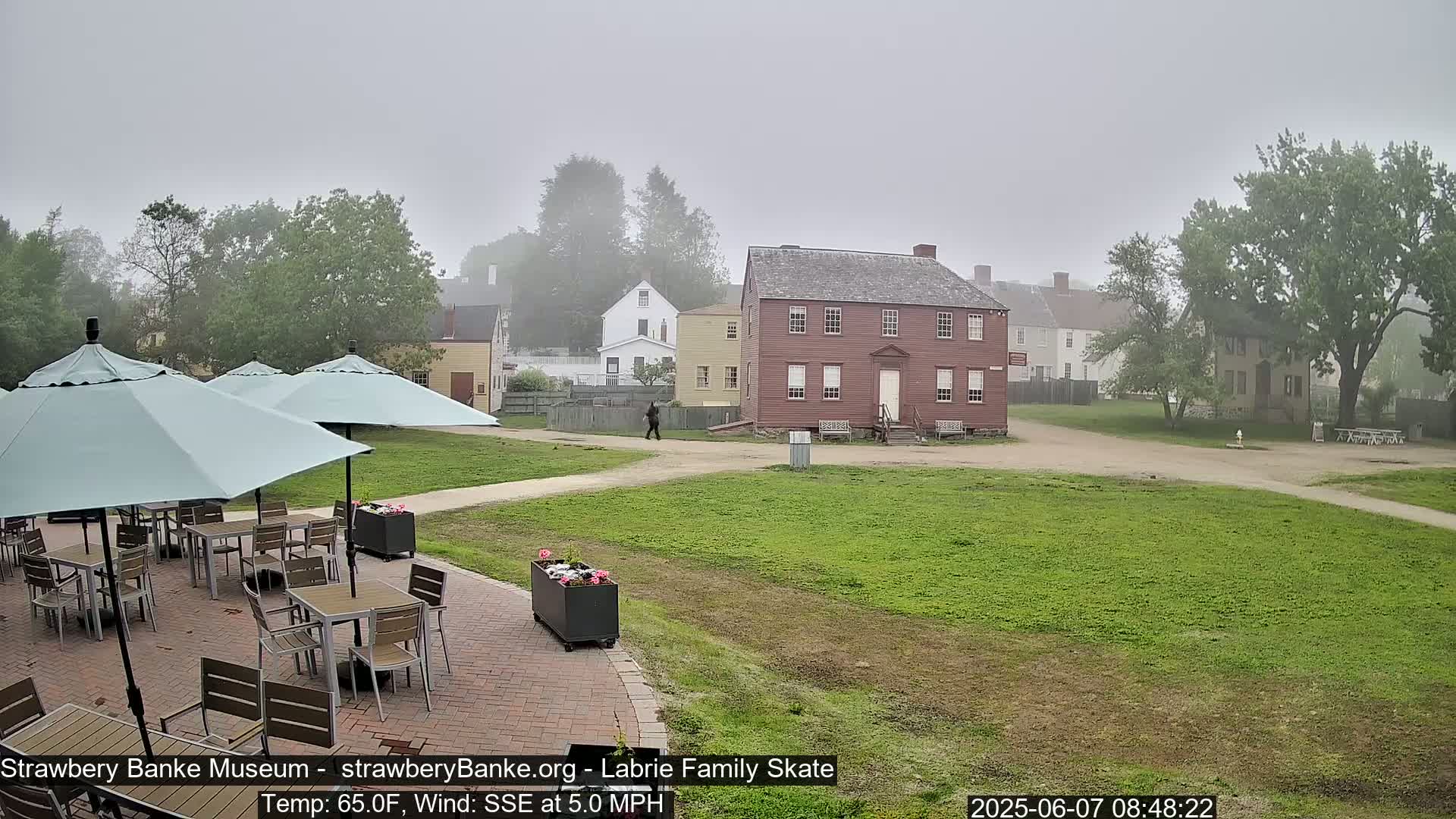 A patio with several tables and teal umbrellas overlooks a grassy area and several historic buildings in a foggy setting.