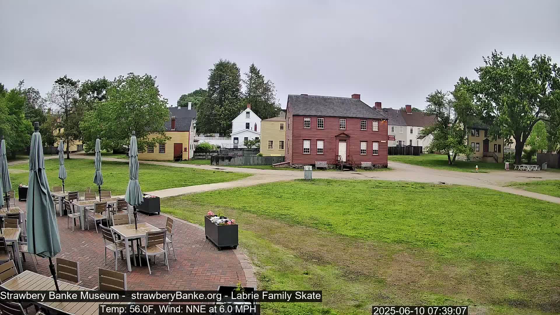 An outdoor patio with tables and umbrellas sits in front of a grassy area and several historic-looking buildings under an overcast sky.