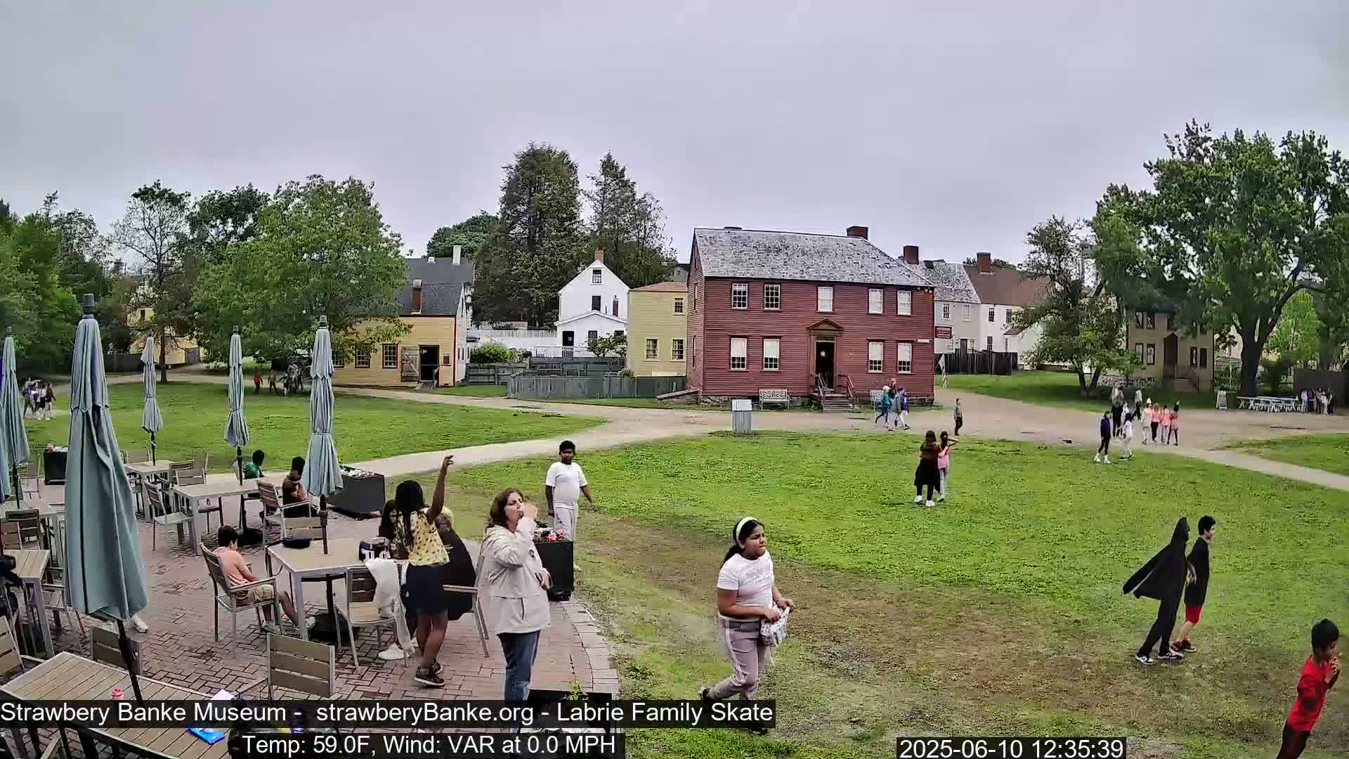 A grassy area with several historical-looking buildings is shown on an overcast day, where people, some children, are walking and sitting at outdoor tables.