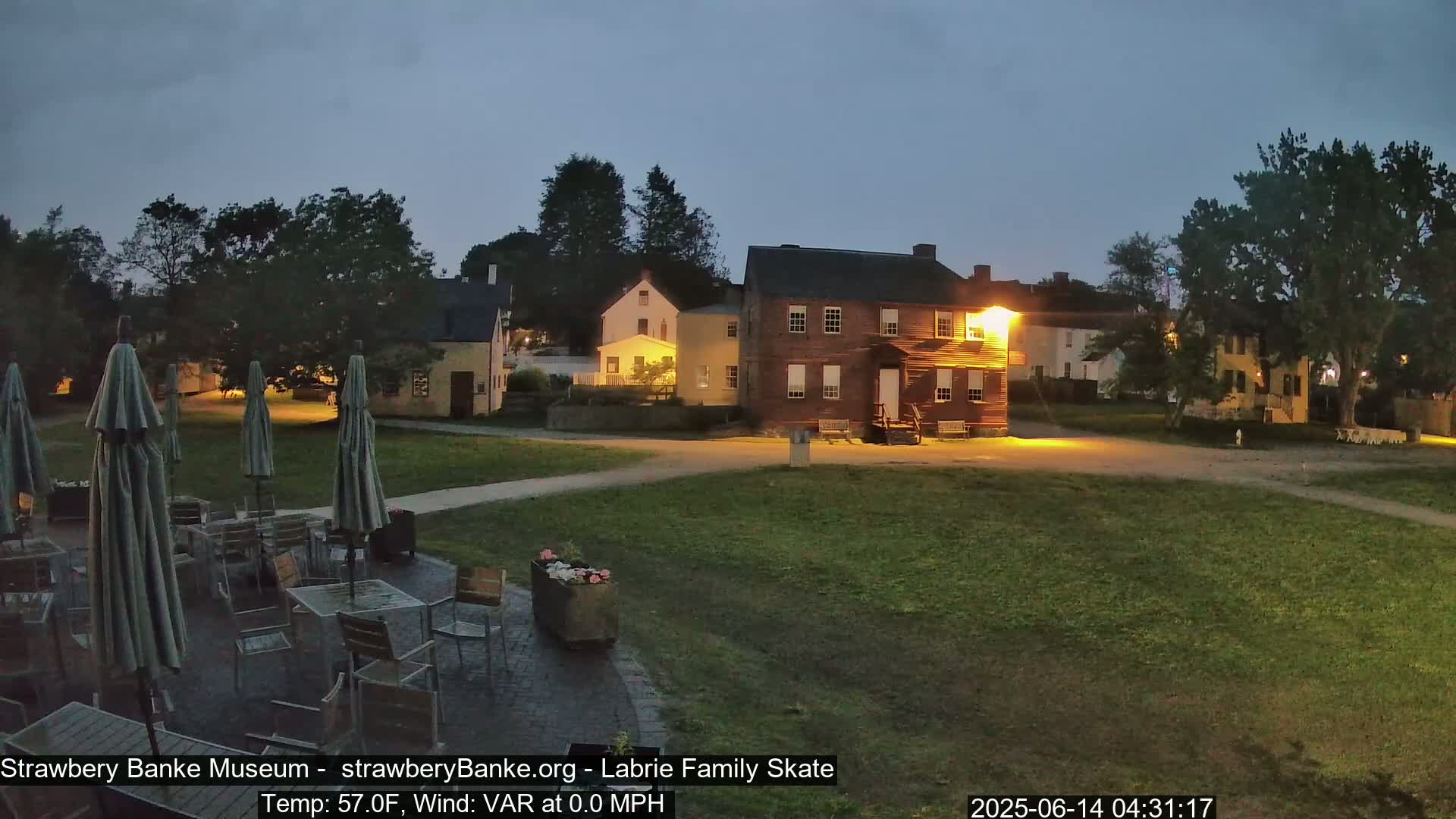 An outdoor scene at twilight shows several historic buildings illuminated, with a patio set furnished with tables and umbrellas in the foreground.