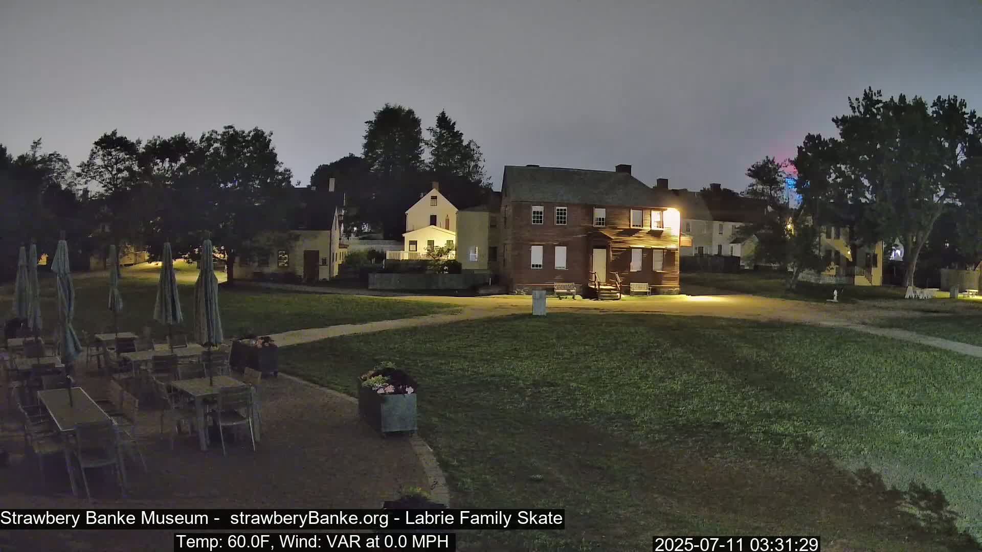 A nighttime scene shows several historical-looking buildings illuminated, a grassy area with patio furniture under closed umbrellas, and trees under a cloudy sky.