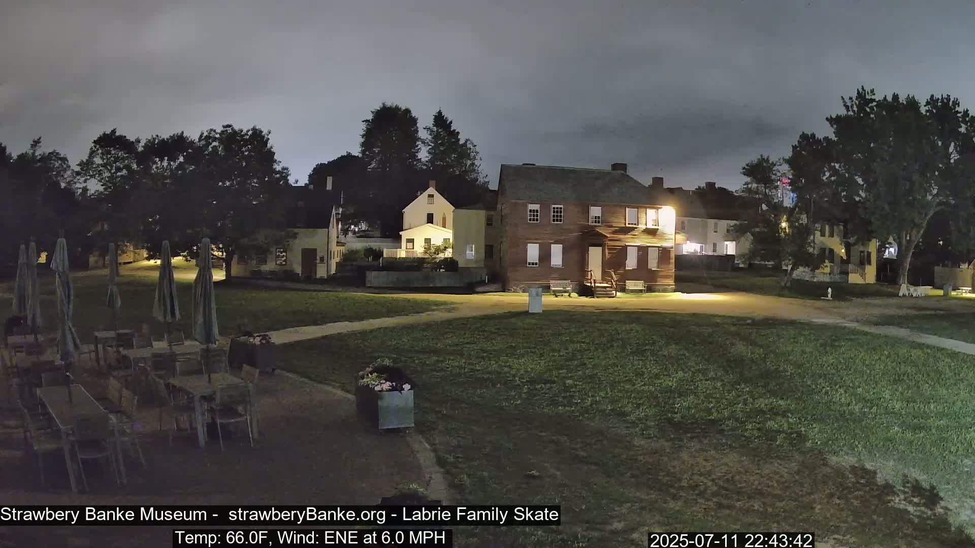 A nighttime scene shows several historical-looking buildings illuminated, a grassy area with patio furniture under closed umbrellas, and trees under a cloudy sky.