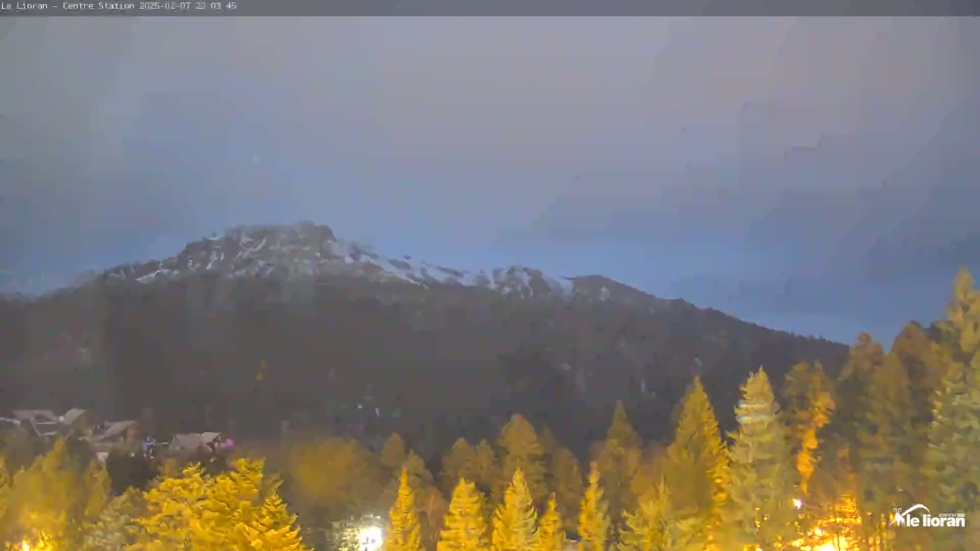 A hazy, dusky sky overlooks a snow-dusted mountain range behind a foreground of illuminated evergreen trees.