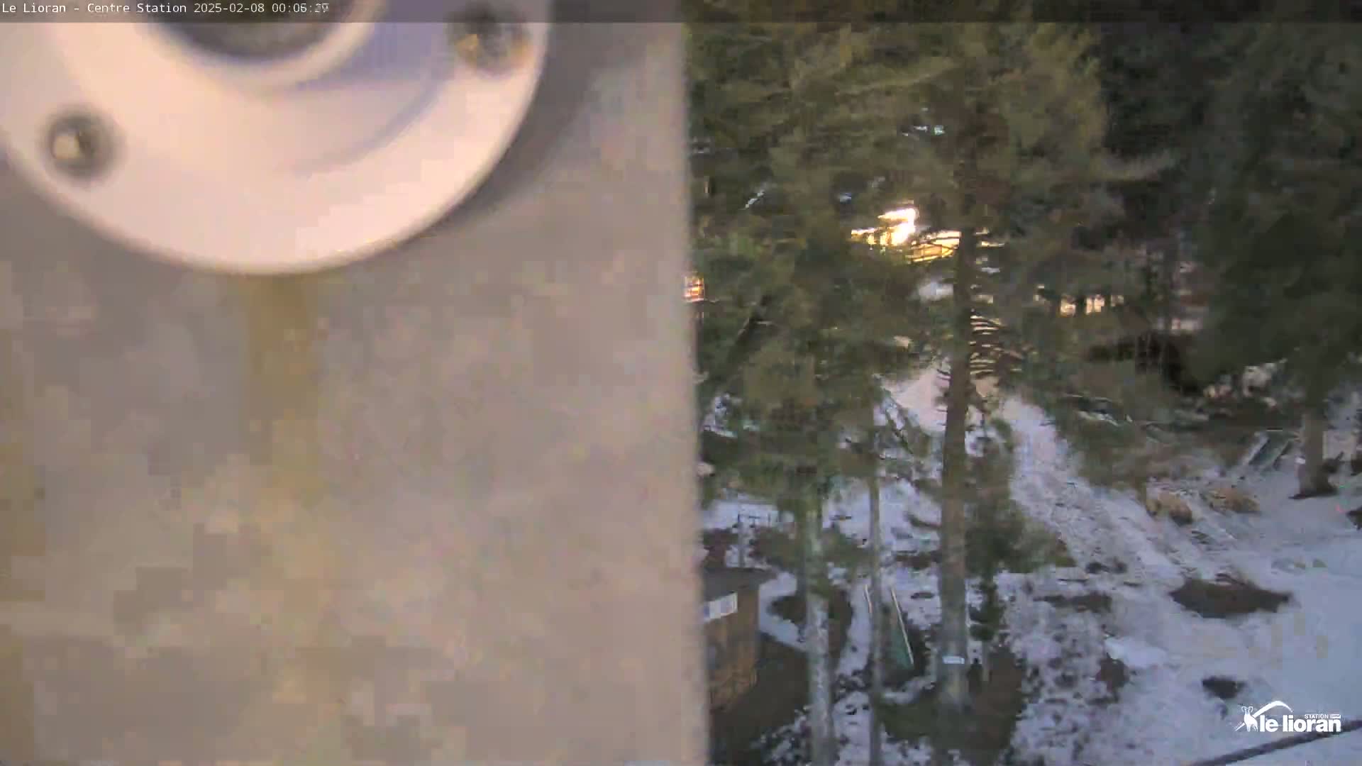 A nighttime view of a snow-covered area with evergreen trees, partially obscured by a light-colored wall and a security camera.