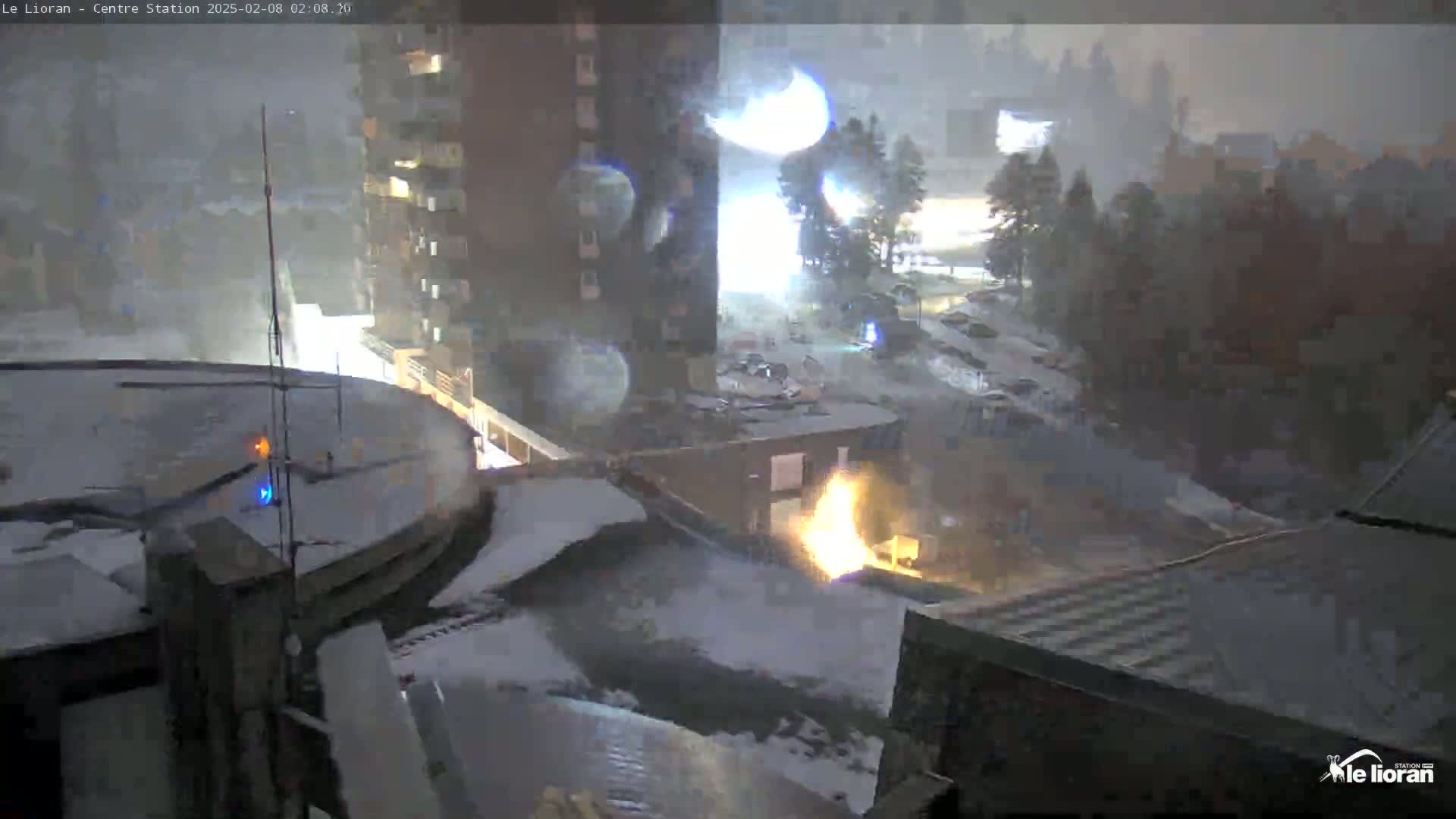 A nighttime scene of a snow-covered town with buildings and a fire visible through heavy snowfall.