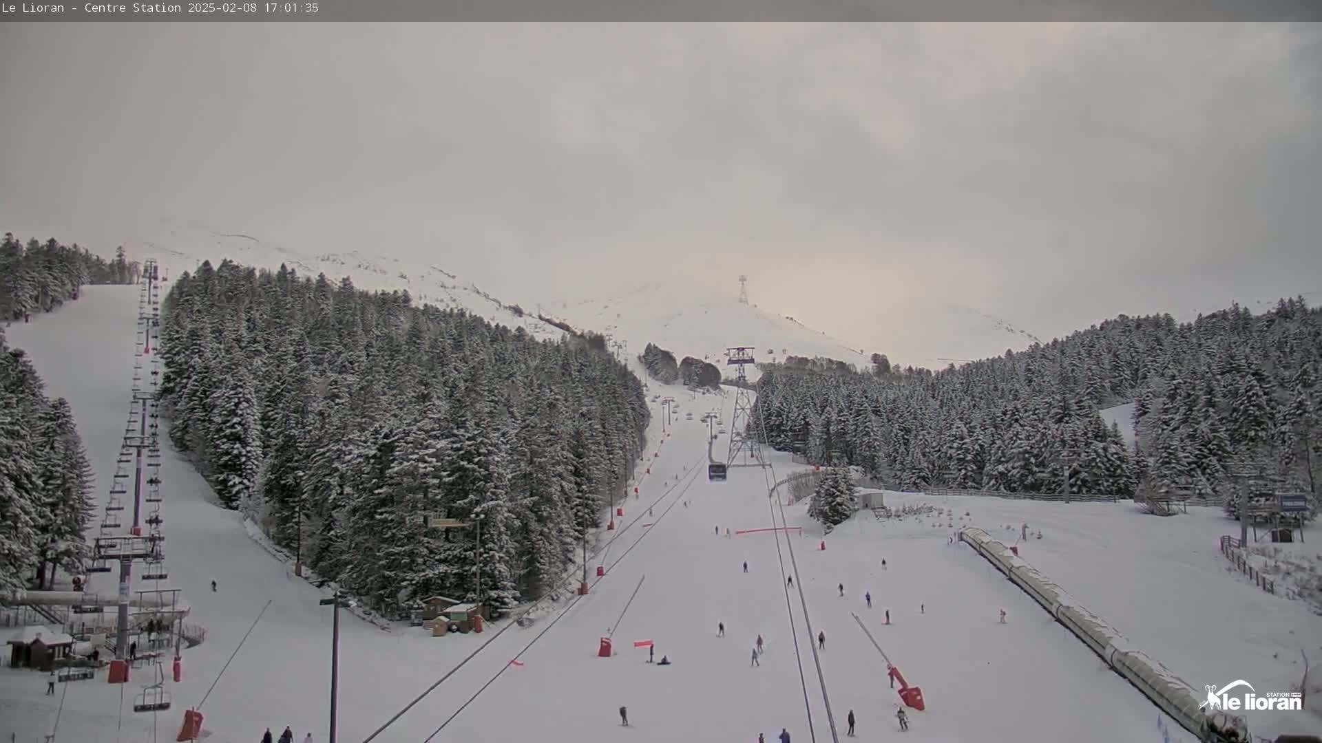 A snow-covered ski slope with several skiers and a chairlift, surrounded by snow-covered evergreen trees under an overcast sky.