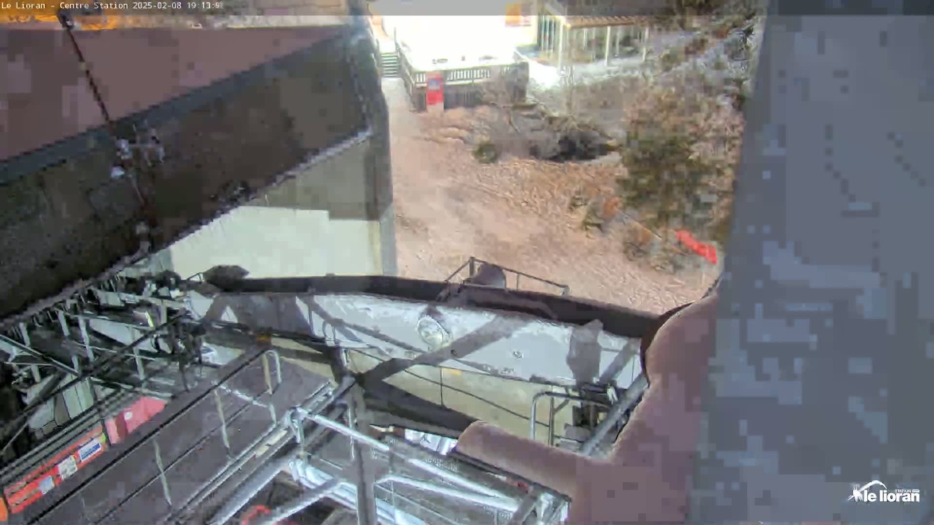 A view from inside a ski lift shows a brown dirt path below with some snow and sparse vegetation, as seen on an overcast day.