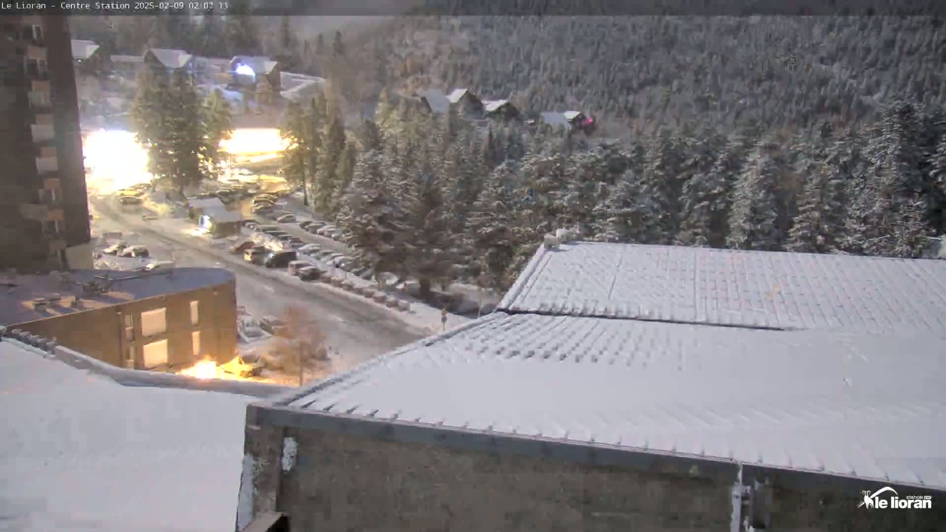 A snow-covered town scene at night, viewed from a rooftop, shows a parking lot, buildings, and a road.
