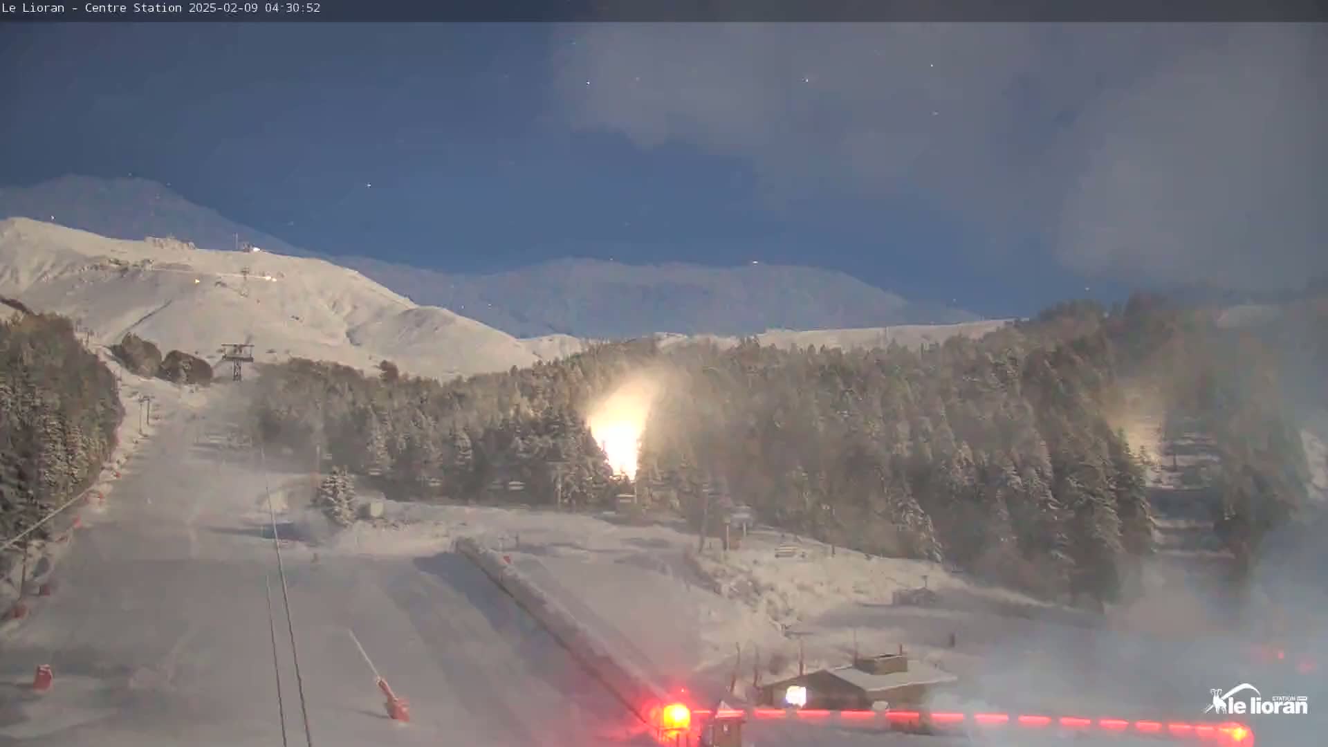 A snow-covered ski slope at night with a snow-making machine actively producing snow, under a clear, dark blue sky.