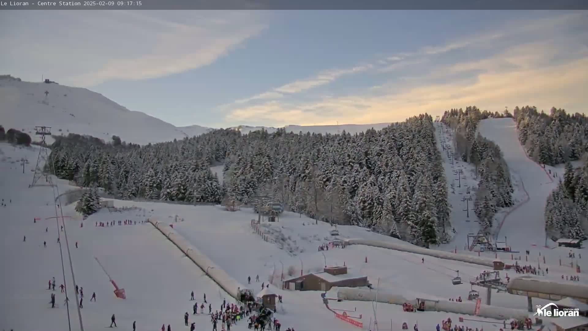 A snow-covered ski resort with skiers and snowboarders on the slopes under a partly cloudy sky.
