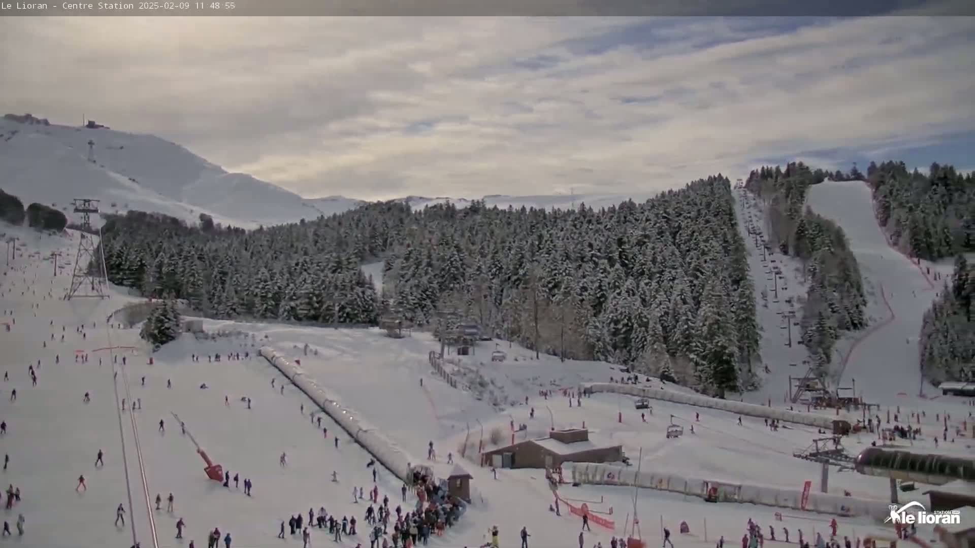 A snow-covered ski resort with skiers and snowboarders on the slopes under a partly cloudy sky.