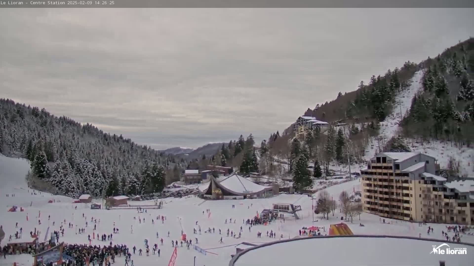 A snow-covered ski resort with several buildings and many people skiing and gathering in a large, open area, under an overcast sky.