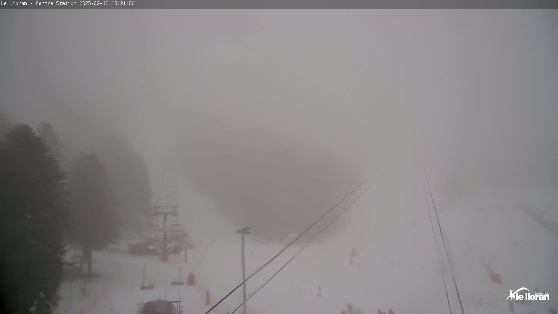 A snow-covered ski slope is obscured by heavy fog, with only parts of the ski lift and surrounding trees vaguely visible.