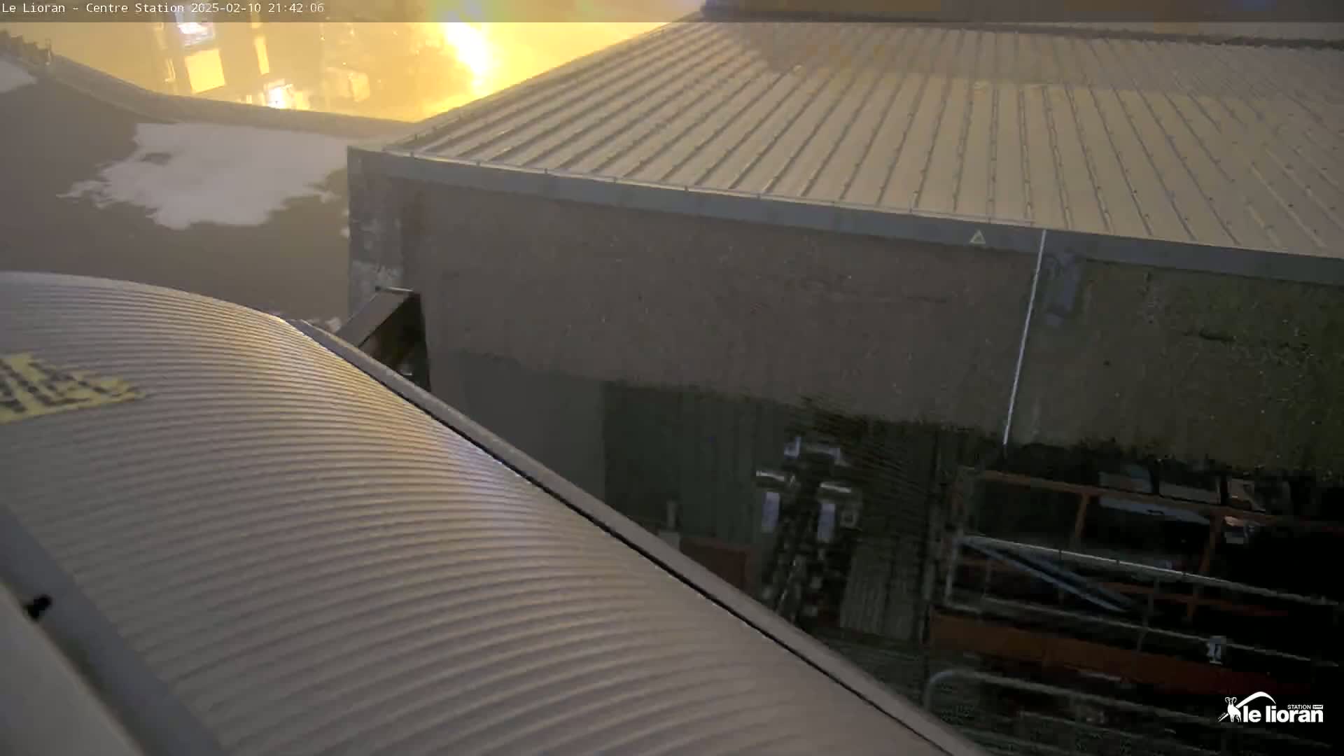 A corrugated metal surface is angled toward a building with a corrugated metal roof, under a dark, possibly rainy sky.