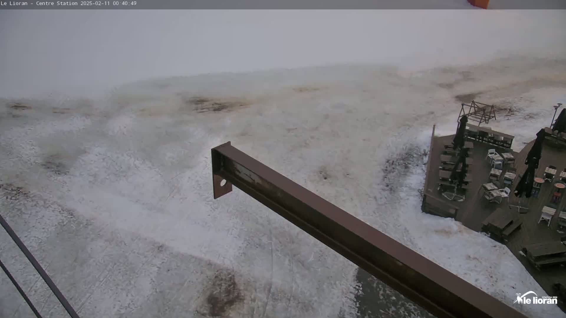 A snowy, foggy scene shows a metal beam extending from the bottom right corner over a snow-covered area, with a partially visible deck and outdoor furniture in the background.