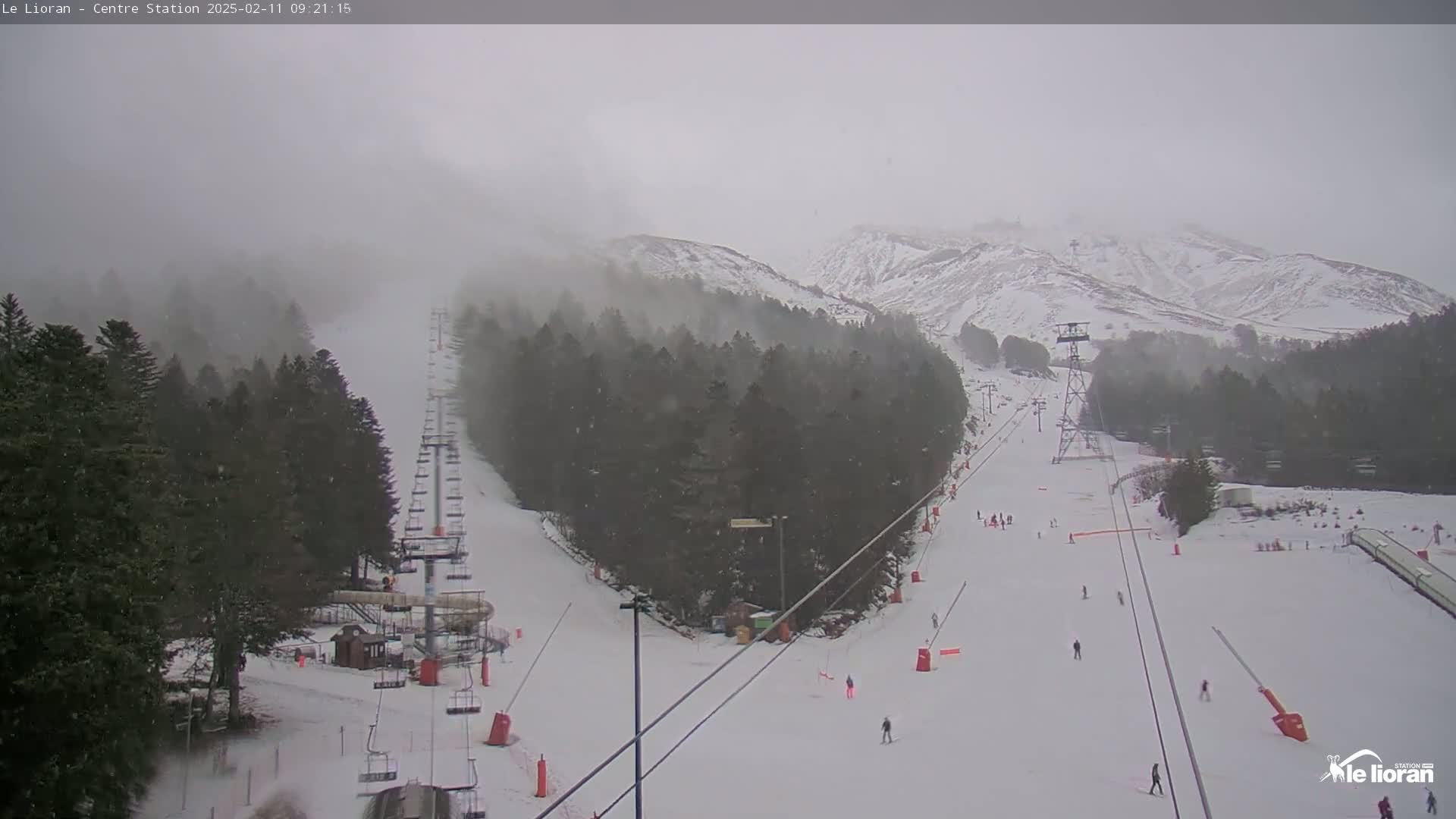 A snow-covered ski slope with a chairlift and several skiers is visible under foggy, snowy conditions, with snow-covered mountains in the background.