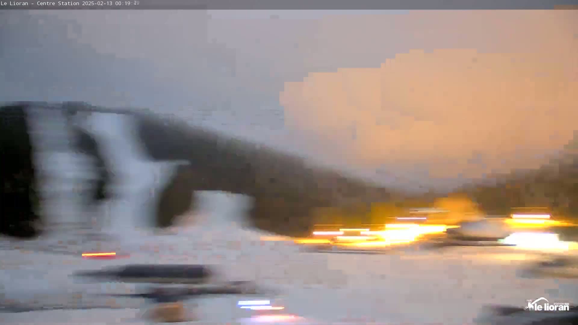A blurry view of a snowy landscape at dusk shows a hill with what appears to be ice formations, along with blurred lights from vehicles in the lower portion of the scene.