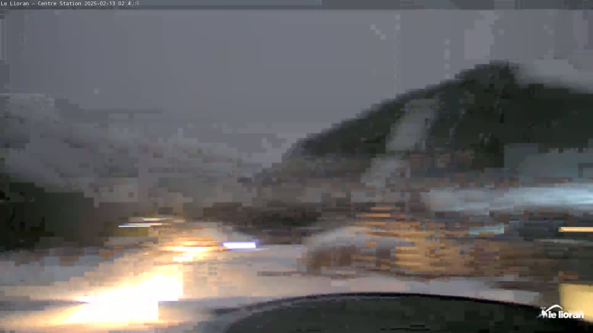 A nighttime, snowy scene is viewed from inside a vehicle, showing blurry headlights and dark buildings in the distance.