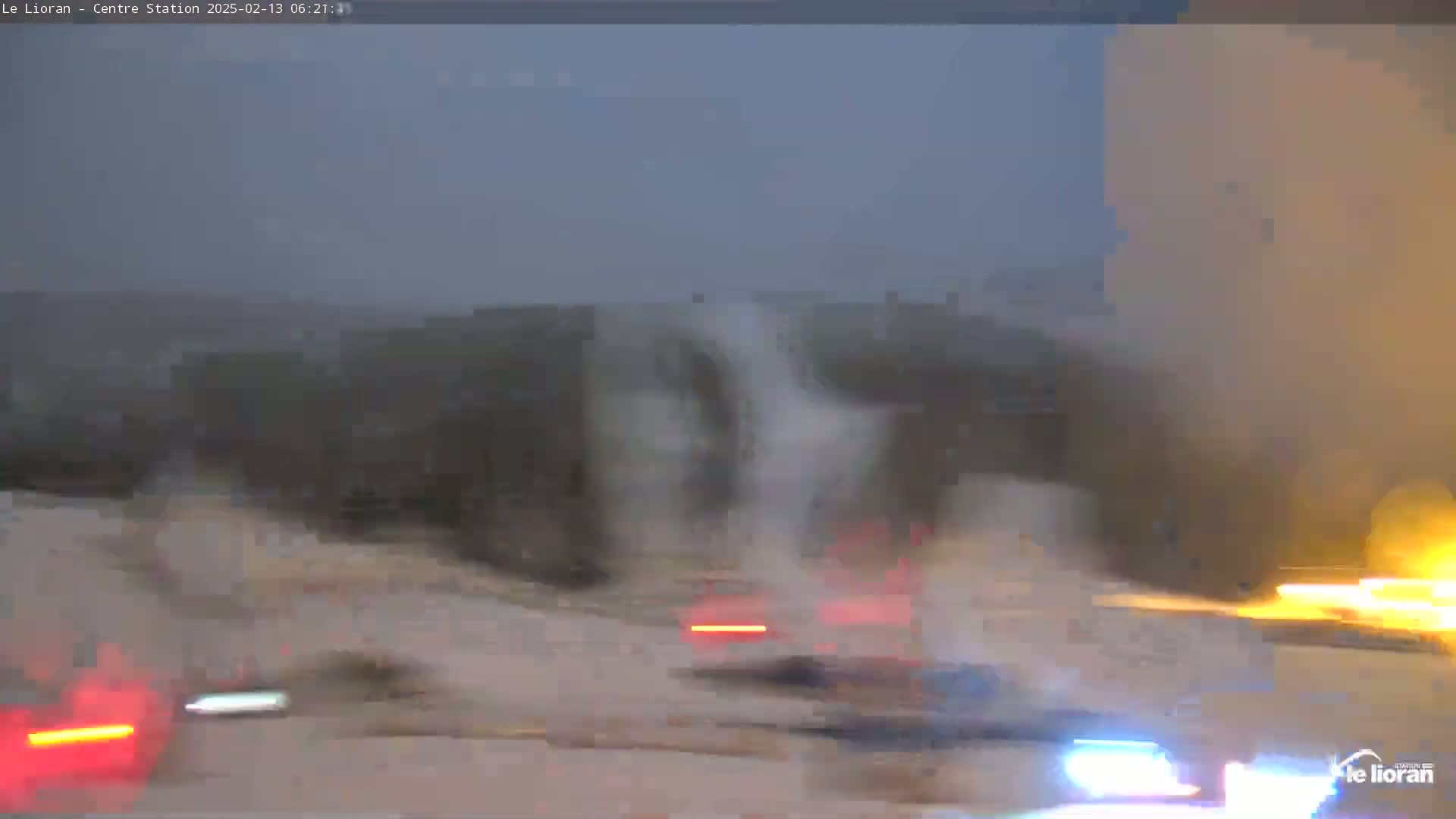 A blurry view of a snow-covered road with several cars driving in the midst of a light snowfall at twilight.