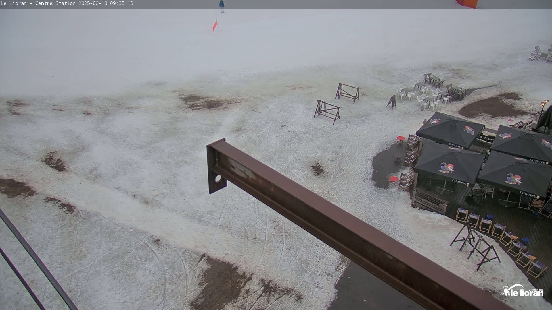 A snow-covered ground with patches of bare earth, several outdoor patio umbrellas and chairs, and a rusty metal beam in the foreground under an overcast sky.