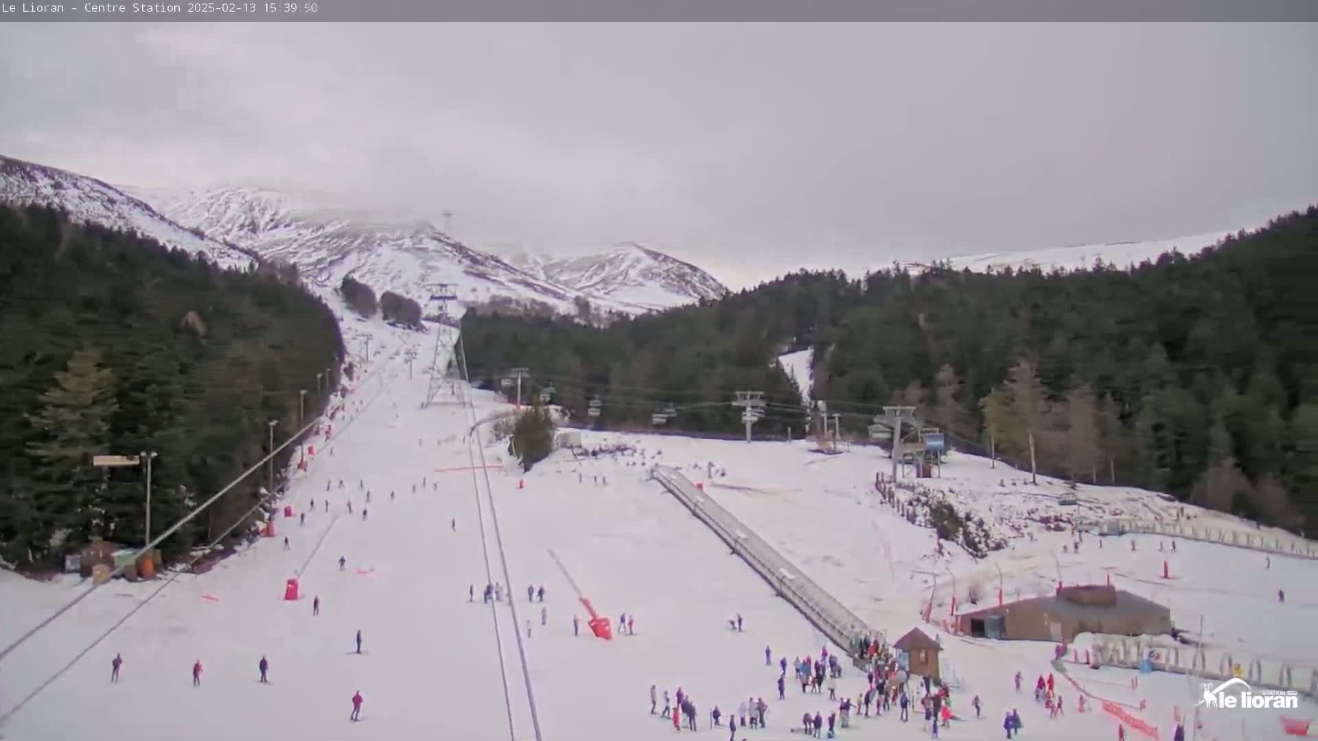 A ski slope with several skiers and snowboarders is visible, under an overcast sky, with snow-covered mountains in the background.