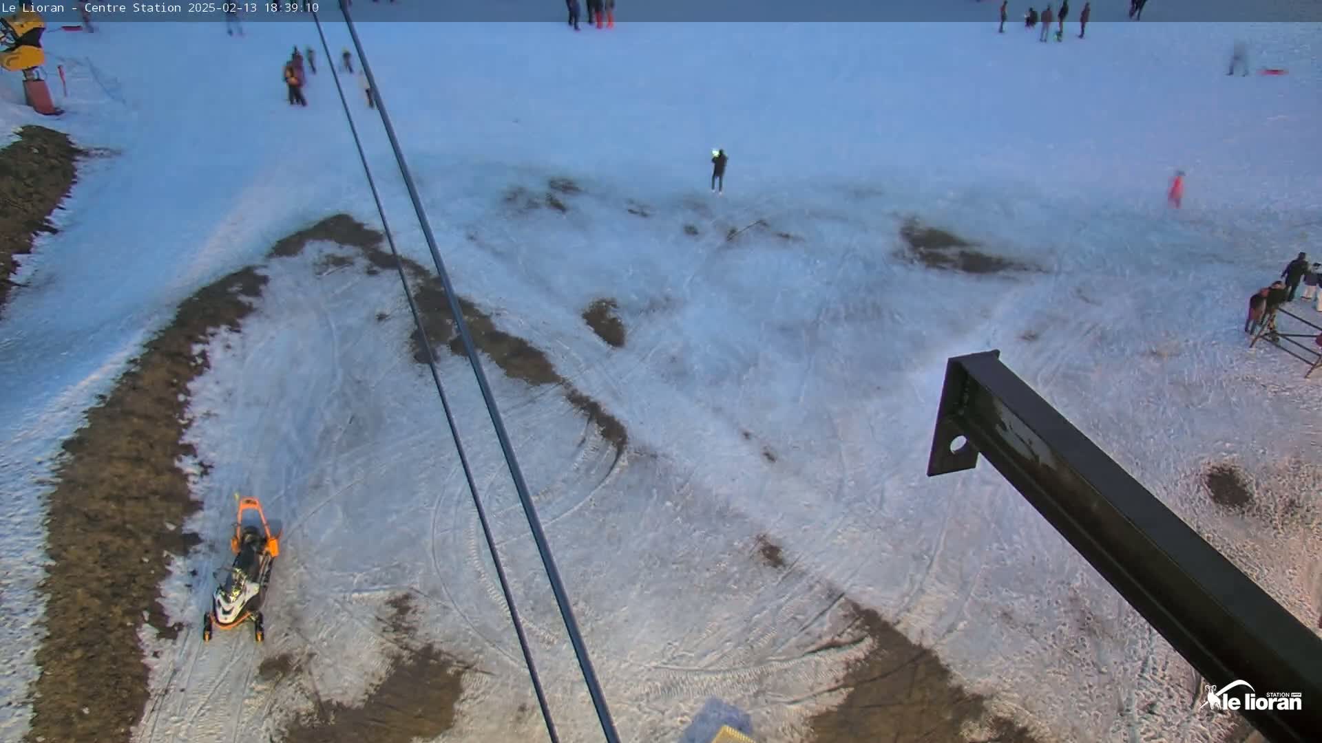 A snow-covered area with patches of brown ground, tire tracks, and a few people scattered about, viewed from a ski lift on a partly cloudy day.