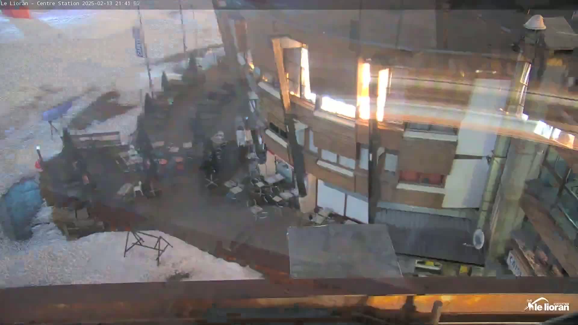 A nighttime high-angle view shows a snow-covered ground outside a multi-story building, with some outdoor furniture and people visible.