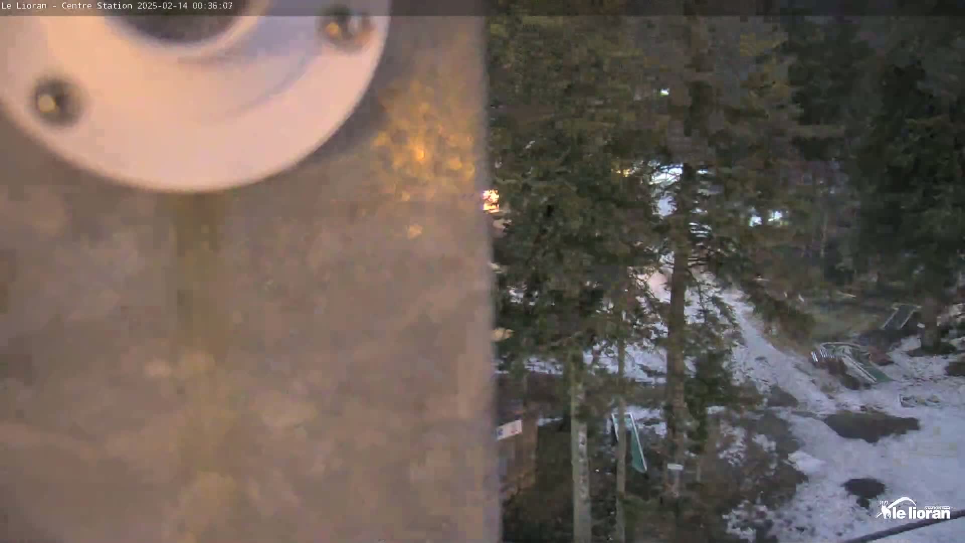 A nighttime view from a security camera shows a snow-covered area with evergreen trees, some equipment, and a portion of a metallic pole dominating the foreground.