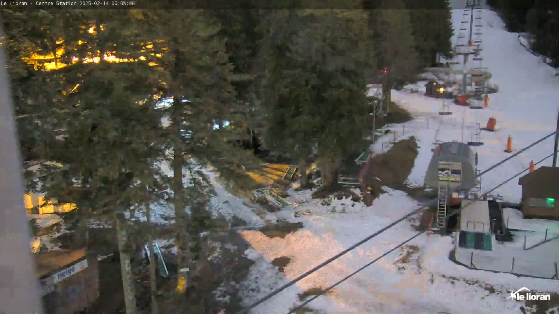A snow-covered ski resort at night, illuminated by lights from buildings, with a chairlift visible in the background.