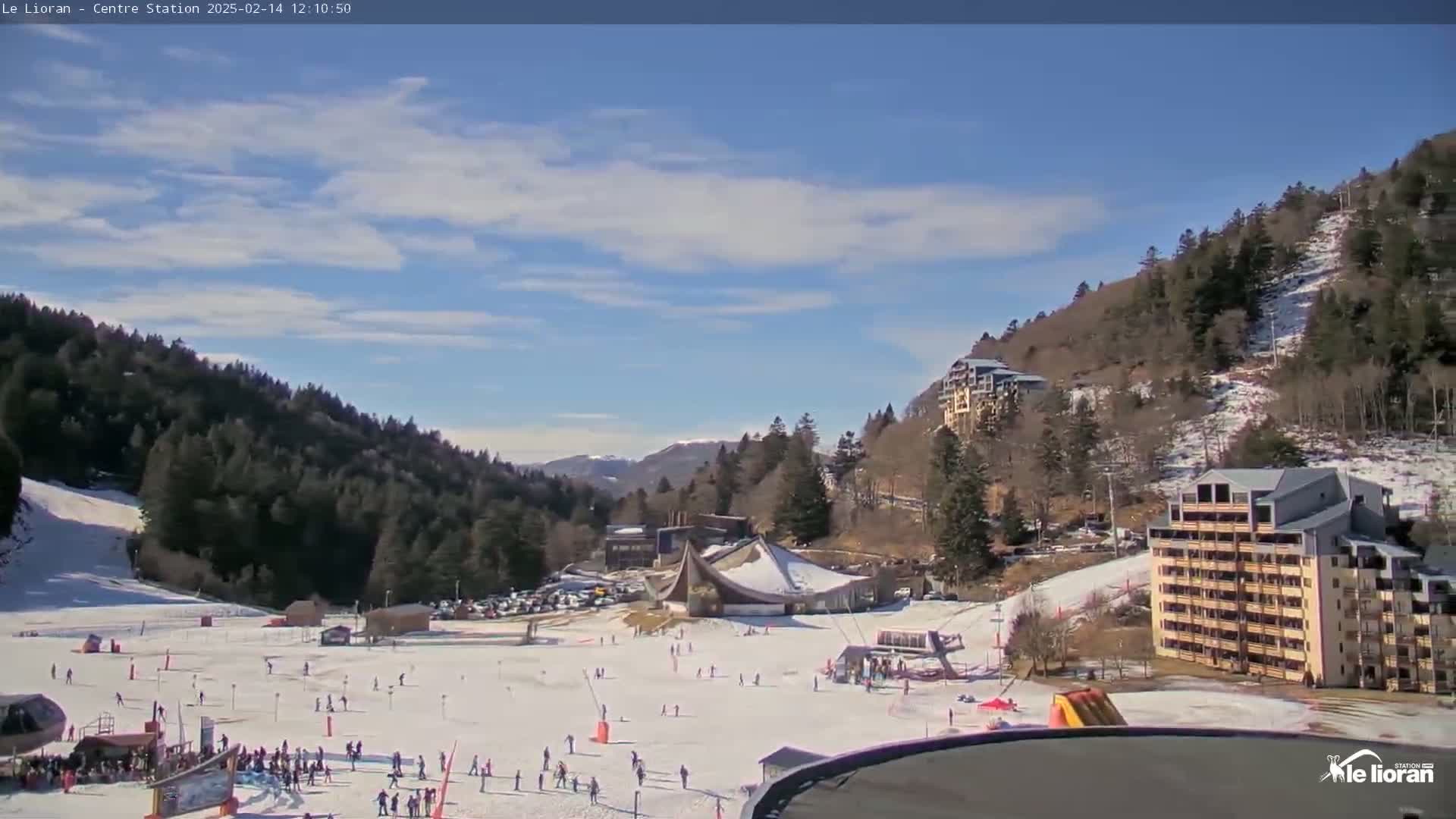 A snow-covered ski resort with several buildings and scattered skiers is visible under a mostly sunny sky with some scattered clouds.