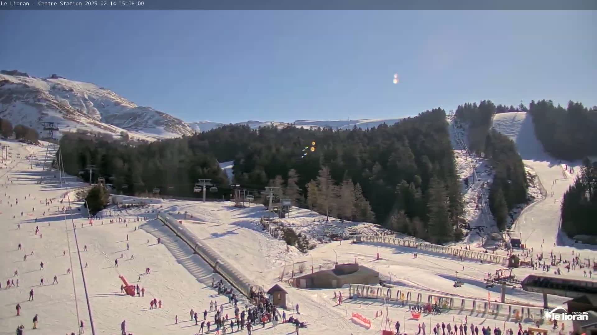 A sunny day at a snow-covered ski resort shows numerous skiers and snowboarders on various slopes, with ski lifts and snow-covered trees in the background.