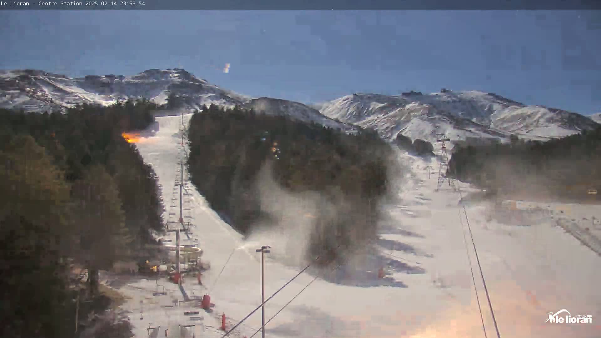 A snow-covered ski slope with a chairlift and snowmaking machines under a clear, twilight sky, set against a backdrop of snow-capped mountains.