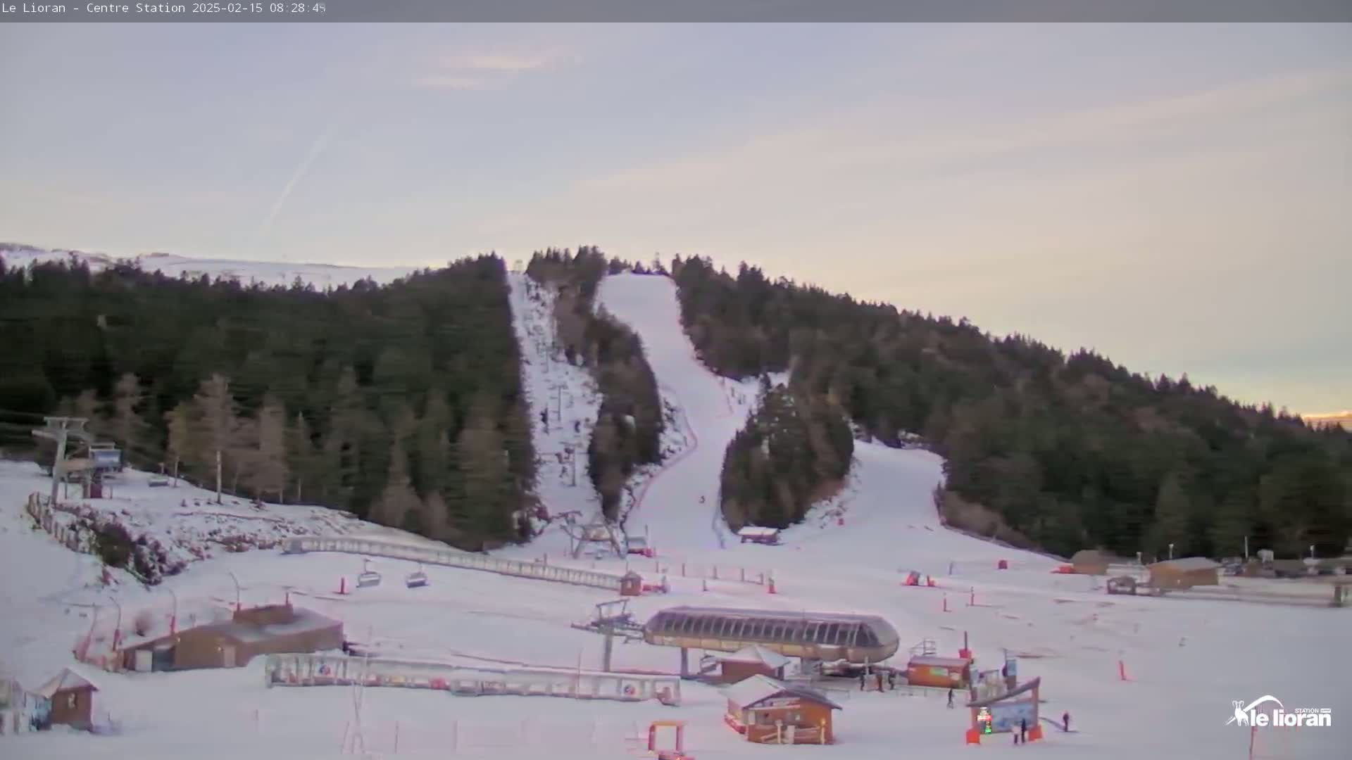 A snow-covered ski slope with several ski lifts and a few skiers is visible under a pale, mostly clear sky.