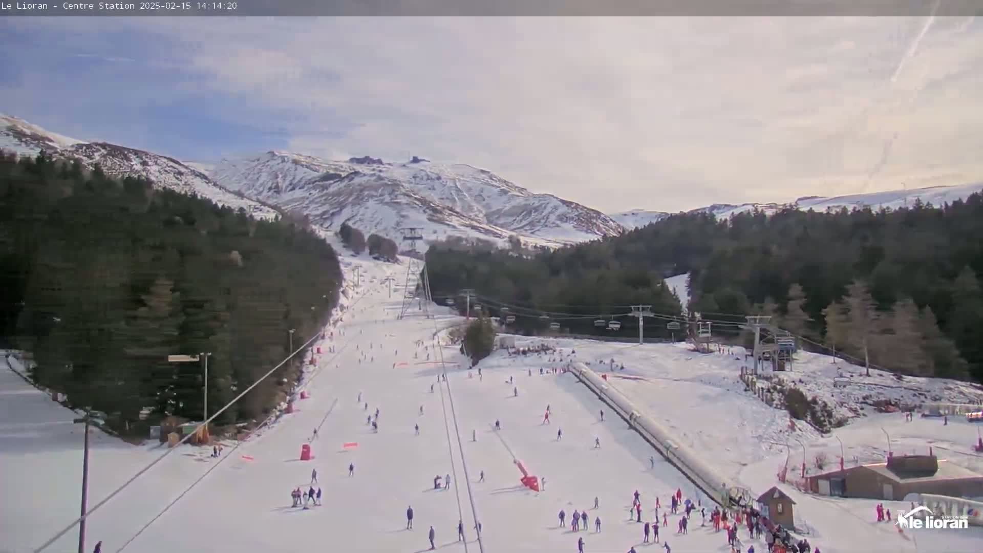 A snowy ski slope with several skiers and snowboarders, ski lifts, and evergreen trees on a partly cloudy day, is nestled in a valley between snow-covered mountains.