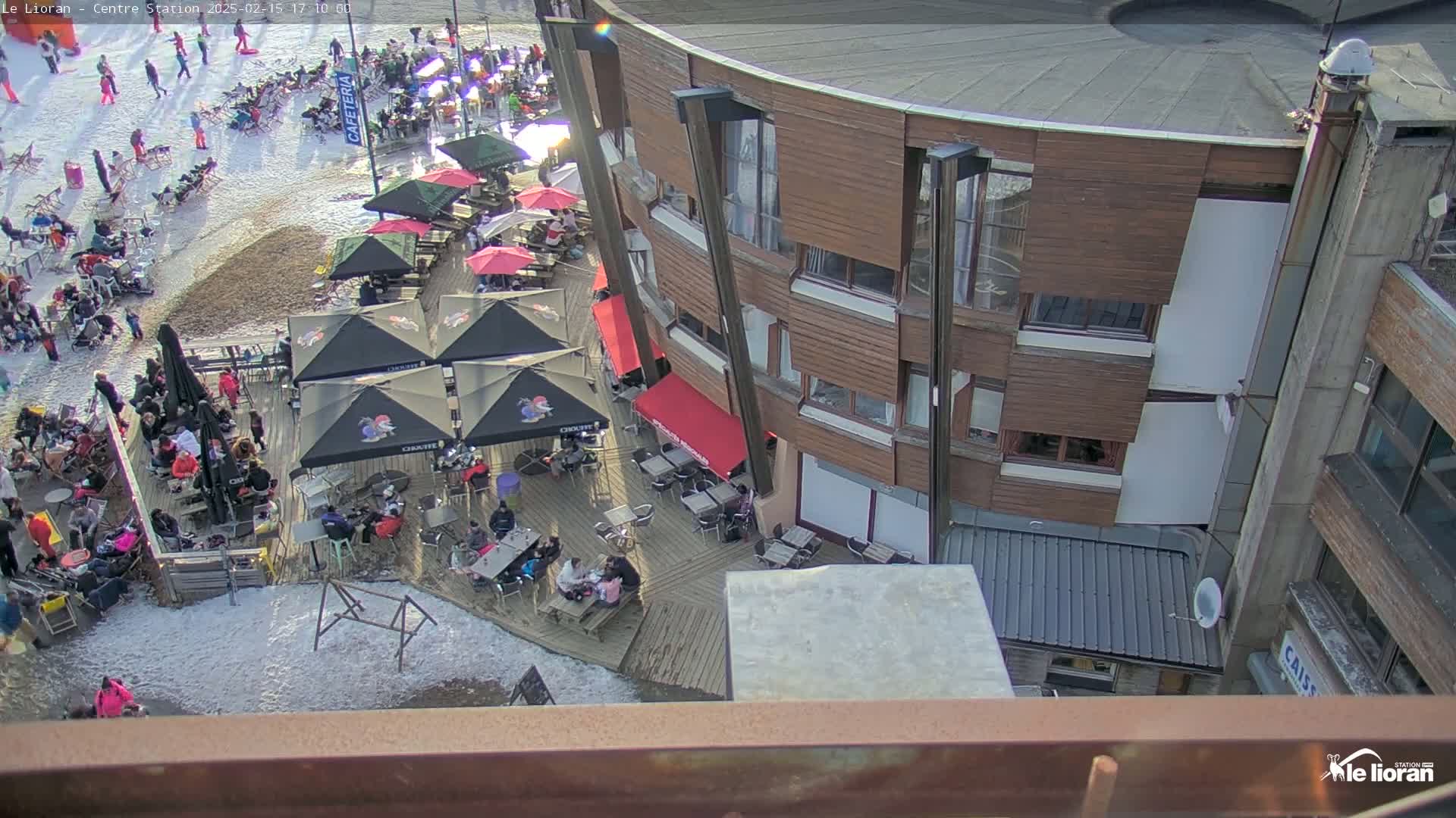 A partially snow-covered outdoor patio area with numerous tables and umbrellas surrounding a multi-story wooden building, under a mostly sunny sky.